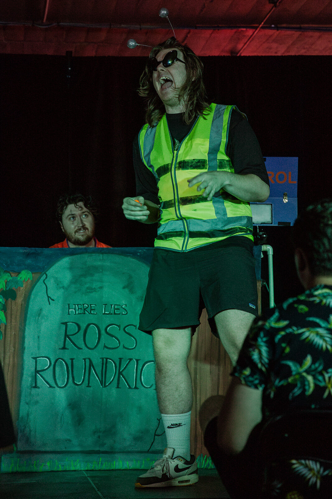 A long-haired performer screaming in a homemade wasp costume in front of a painted gravestone, with Karate Man peering out from behind it